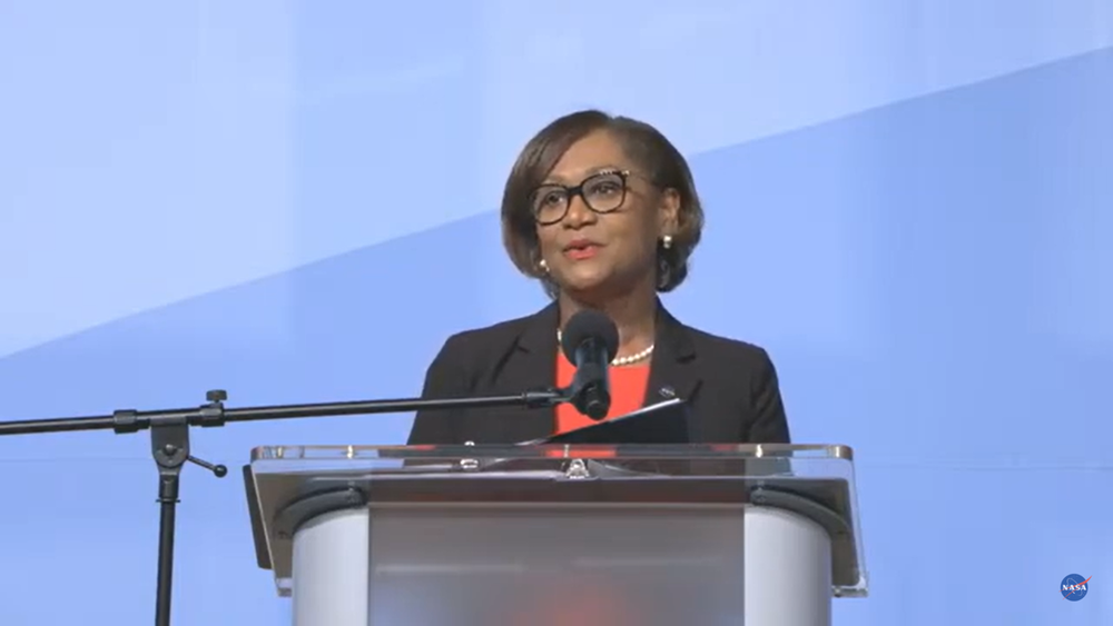 A Black woman with short hair and glasses stands behind a podium, delivering remarks.